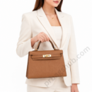 brown-kelly-style-top-handle-handbag-held-by-woman-in-white-blazer-on-white-background
